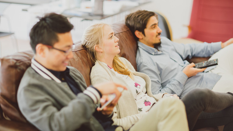 Three students, two male and one female, sit together on a sofa in their flat watching tv